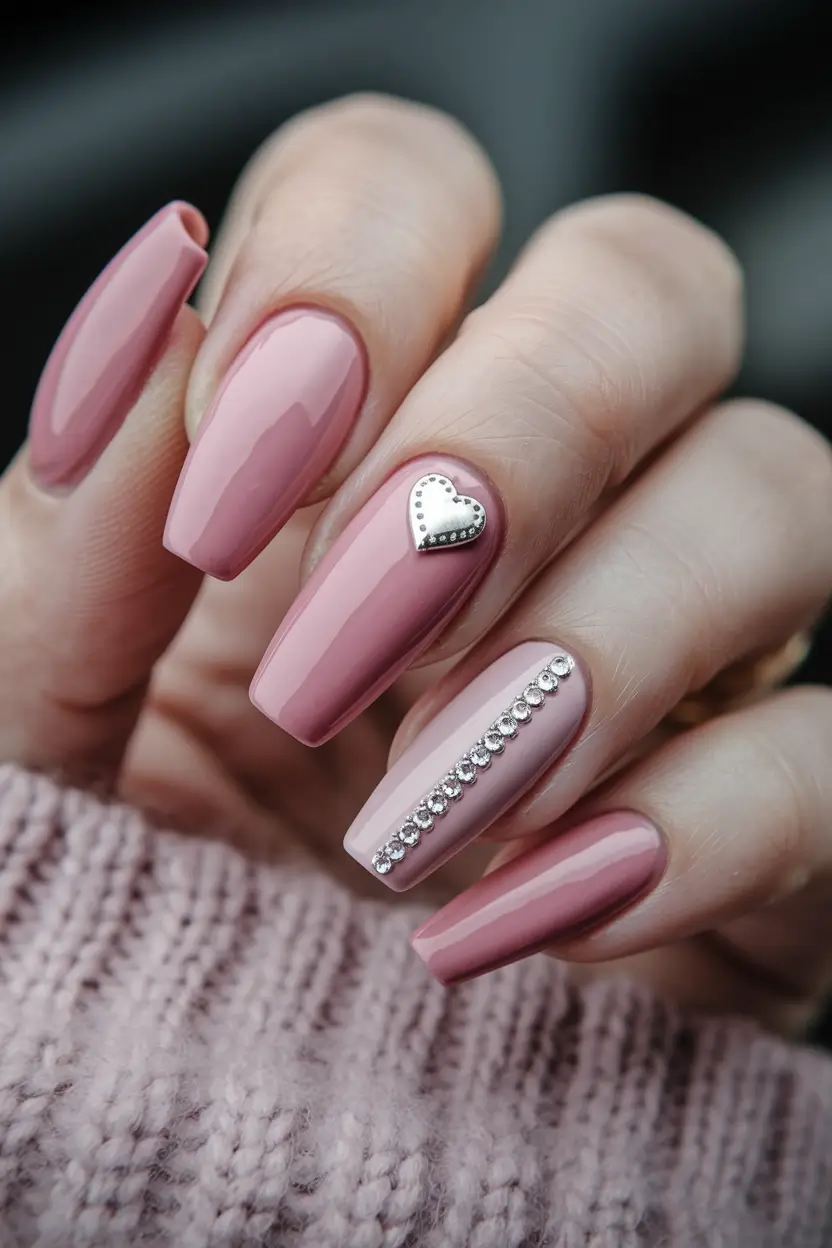 A manicure with rosy mauve polish, tiny rhinestones, and a heart charm on fair skin.
