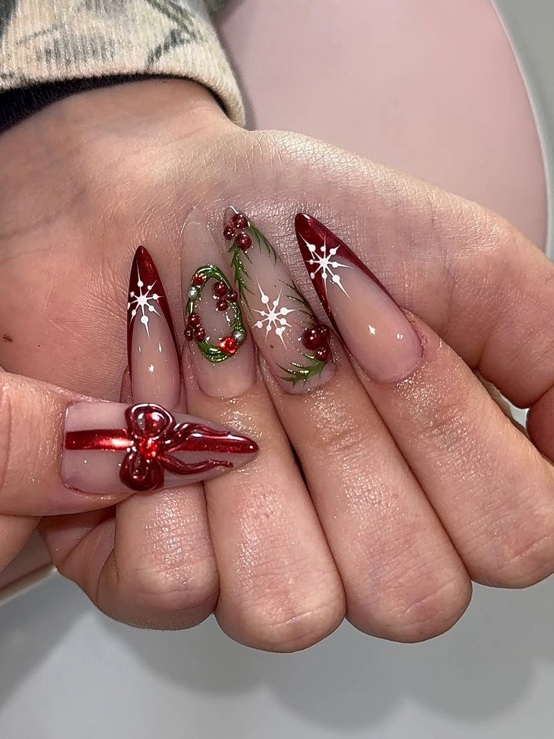 Long pointed nude nails with red tips, wreaths, snowflakes, and raised bow