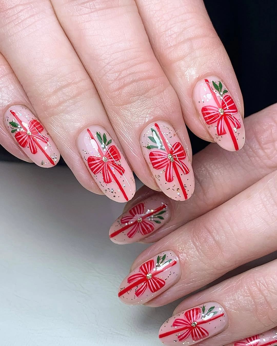 Almond blush nails with red ribbon bows, green sprigs, and fine speckles